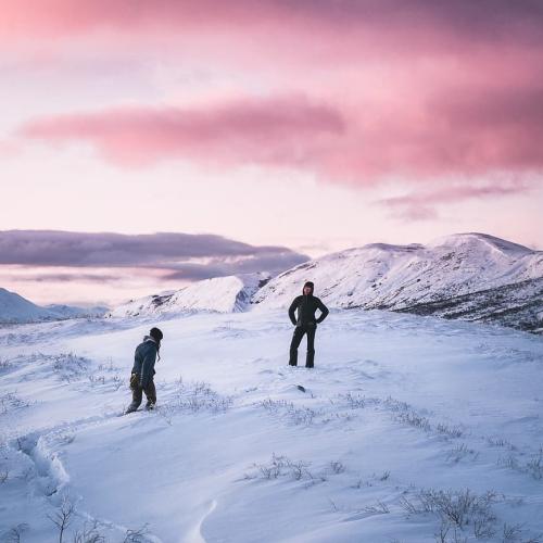 Two hikers walk across a snowy mountain plateau under a pink sky